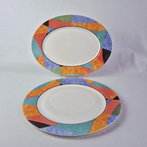 Puede incluir: Dos fuentes ovaladas. Los platos tienen un centro blanco con un borde negro. El borde exterior presenta un diseño geométrico colorido con formas naranjas, rojas, azules, verdes y negras. Los platos se muestran sobre un fondo blanco.