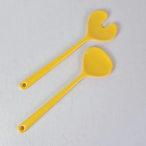 May include: Two bright yellow salad servers. One server has a spoon-shaped head, and the other has a fork-shaped head. Both servers have long, slender handles with a hole at the end for hanging. The servers are made of a smooth, glossy material.