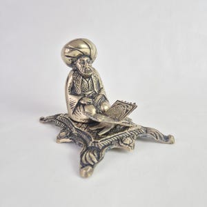 Könnte beinhalten: Messingfigur einer sitzenden Person mit Turban, die ein Buch liest. Die Figur steht auf einem dekorativen Sockel mit aufwendigen Mustern. Die Farbe ist ein gedämpftes Gold. Die Figur ist wahrscheinlich aus Messing.