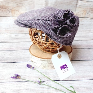 Puede incluir: Una gorra de tweed de color morado y blanco con un gran adorno de flor morada. La gorra se muestra en un soporte de madera con una etiqueta blanca que dice "Bunny Hill Baby & Kids".