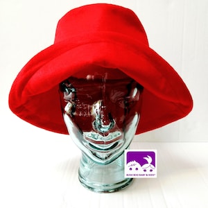 May include: A bright red bucket hat with a wide, downward-sloping brim. The hat is displayed on a clear, translucent mannequin head. A small square logo featuring two stylized faces and the text BUSH BOO BABY & KIDS is visible below.