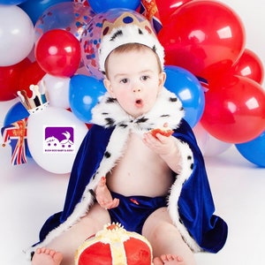May include: A baby wearing a blue velvet king costume with a white fur trim and a gold crown sits in front of a red, white and blue balloon arch. The baby is holding a red cupcake and looking at the camera. The baby is sitting in front of a red and gold cake shaped like a crown.