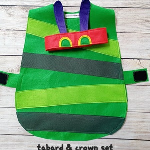 Peut inclure: Ensemble tabard et couronne en feutre vert sur le thème de la chenille. Le tabard présente des rayures horizontales de différentes nuances de vert et de gris. Un bandeau rouge avec des accents jaunes et verts est posé dessus. Le texte "tabard & crown set" est en bas.