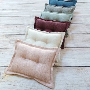 May include: A set of seven decorative pillows in various colors and textures, including teal, gray, green, burgundy, beige, and pink. The pillows are arranged in a row on a light wood surface.
