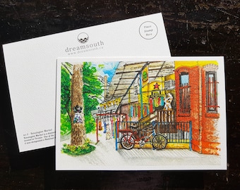 May include: A watercolor illustration of Kensington Market in Toronto, Canada. The illustration features a colorful building with a red star and a sign that says "Kensington Market". There are clothes hanging on a line outside the building, and a bicycle is parked in front of it.