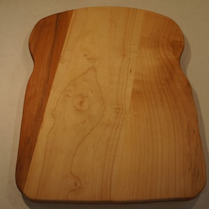 May include: A wooden cutting board in the shape of a slice of bread. The board is made of light-colored wood with a darker brown section on the left side. The wood grain is visible.
