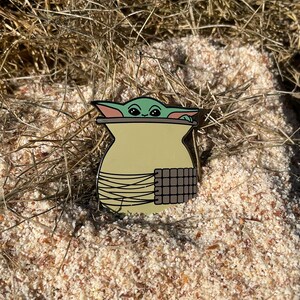 Puede incluir: Un pin de esmalte con un personaje de la franquicia Star Wars, conocido como "The Child" o "Baby Yoda", asomándose desde una cesta tejida. El pin es verde y marrón.