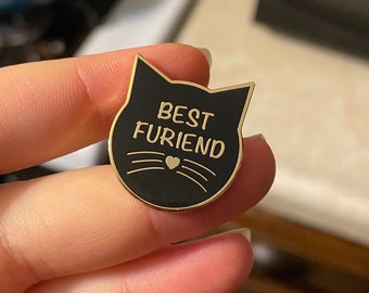 May include: Black cat-shaped enamel pin with gold trim. The pin says "Best Furriend" with a small heart in the middle.