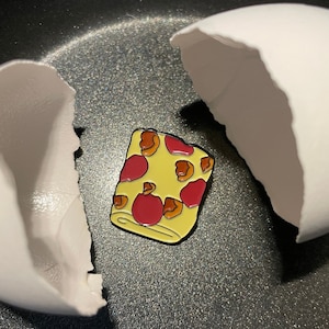 May include: A yellow and red enamel pin shaped like a pizza slice with a white background. The pin is sitting inside of a broken eggshell.