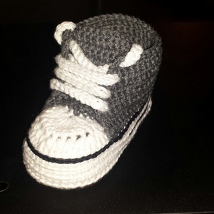 May include: A high-top sneaker bootie crafted from dark gray yarn with a white crocheted toe cap, white laces, and a white sole featuring a thin black horizontal stripe detail. The footwear is constructed with a textured, hand-stitched knit pattern.