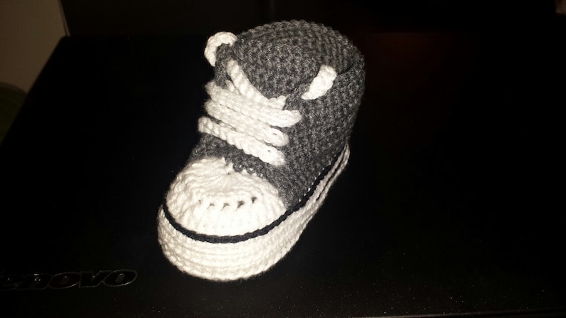 May include: A high-top sneaker bootie crafted from dark gray yarn with a white crocheted toe cap, white laces, and a white sole featuring a thin black horizontal stripe detail. The footwear is constructed with a textured, hand-stitched knit pattern.