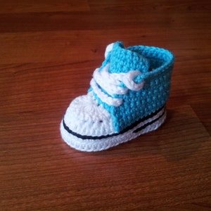 May include: A crochet baby shoe designed to look like a high-top sneaker. The shoe features a bright blue upper section, a white toe cap, white laces, and a white sole with a thin black horizontal stripe running along the side of the base.