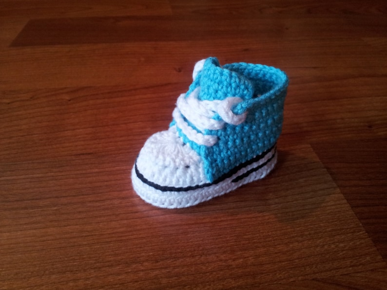 May include: A crochet baby shoe designed to look like a high-top sneaker. The shoe features a bright blue upper section, a white toe cap, white laces, and a white sole with a thin black horizontal stripe running along the side of the base.