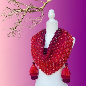 May include: A handmade, red and pink infinity scarf with a textured, scale-like pattern. The scarf is adorned with small, gold-colored beads and has two matching tassel pendants. The scarf is displayed on a white mannequin.