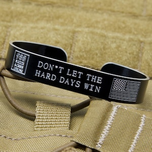 May include: A black metal cuff bracelet with the text "DON'T LET THE HARD DAYS WIN" and an American flag design. The bracelet also features the text "988 Suicide & Crisis Lifeline". The bracelet is set against a tan background.