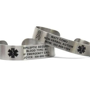 Puede incluir: Dos pulseras de alerta médica de acero inoxidable. La pulsera superior tiene una cinta negra y el texto "MATT COMO NON-VERBAL AUTISM MOM: 303-555-1284". La pulsera inferior tiene un símbolo negro de estrella de la vida y el texto "EPILEPTIC SEIZURES BLOOD TYPE: A+ IF EMERGENCY CALL JOYCE: 208-555-1028".