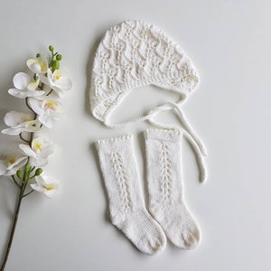 May include: A white knitted baby bonnet with a matching pair of socks. The bonnet has a lacy pattern and ties under the chin. The socks are knee-high and also have a lacy pattern.