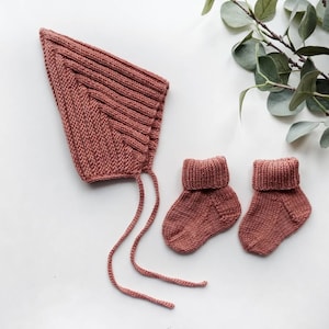 May include: A set of knitted baby accessories in a dusty rose color. The set includes a bonnet with ties and two booties.