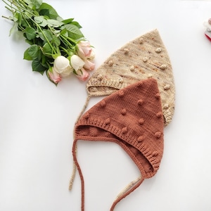 Peut inclure: Deux bonnets de bébé en tricot avec des liens. L'un est brun avec des pompons blancs et l'autre est beige avec des pompons multicolores.
