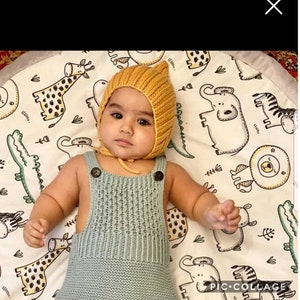 Puede incluir: Un bebé con un mono de punto azul claro y un gorro de punto amarillo. El bebé está tumbado en una alfombra de juego blanca con estampado de animales en blanco y negro.