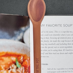 May include: A wooden spoon-shaped bookmark with the text "STIR THE POT" printed on the handle. The bookmark is brown and is placed on a cookbook page with text and a food image.
