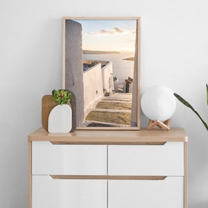 May include: A framed photograph of a white building with steps leading up to a view of the ocean. The photo is on a wooden dresser with a white lamp and a small potted plant.