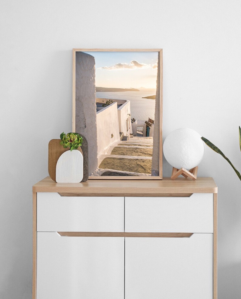 May include: A framed photograph of a white building with steps leading up to a view of the ocean. The photo is on a wooden dresser with a white lamp and a small potted plant.