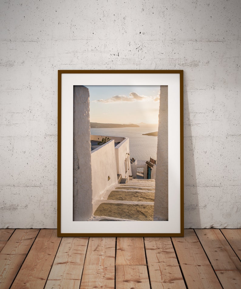 May include: A framed photograph of a white stone staircase leading up to a view of a blue ocean and a small island in the distance. The photo is taken from a narrow alleyway with white walls on either side.