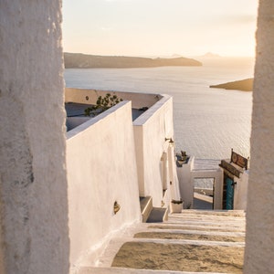 May include: Whitewashed steps leading up to a building with a view of the ocean and a distant island. The steps are made of stone and have a rough texture. The building is white and has a simple design. The ocean is blue and calm. The sky is a light blue and there are some clouds in the distance.
