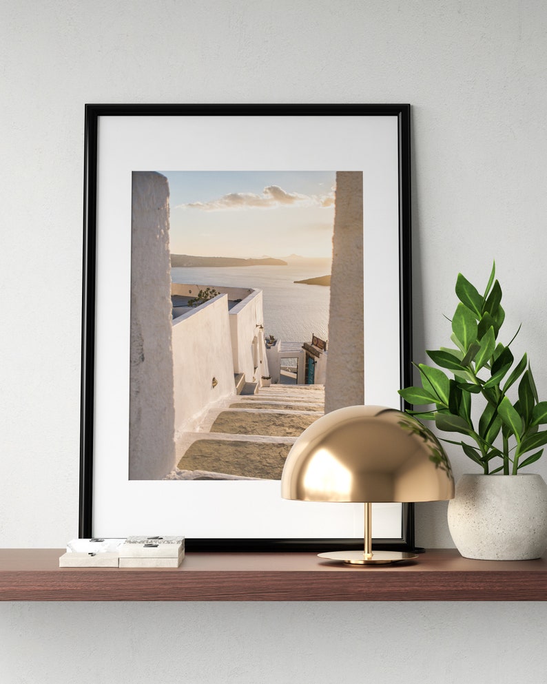 May include: A framed photograph of a white, winding staircase leading to a view of a blue ocean and a small island in the distance. The photo is on a shelf with a gold table lamp and a potted plant.