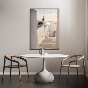 May include: A white round table with a gray concrete base, two wooden chairs, and a framed photograph of a white building with steps leading down to a blue body of water.