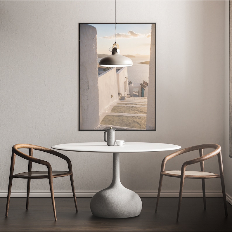 May include: A white round table with a gray concrete base, two wooden chairs, and a framed photograph of a white building with steps leading down to a blue body of water.