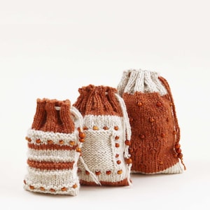 May include: Three hand-knitted drawstring bags in varying sizes. The bags feature a color block design with rust-colored and cream-colored yarn. The bags are adorned with small, amber-colored beads. The bags are ideal for small gifts or storage.