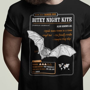 May include: Black t-shirt featuring a white graphic of a bat with the text "Know Your DANGER BOIS BITEY NIGHT KITE (GANGLEE DANGLEE) ALSO KNOWN AS: Upside Down Frown In A Gown Angel Rat Un-friendly Frisbee Vampire Flap Flap FACT FILE DANGER: AAAAA SECOND COUSIN: THE UMBRELLA BPN (BITES PER NIGHT): REF: ROBIN"