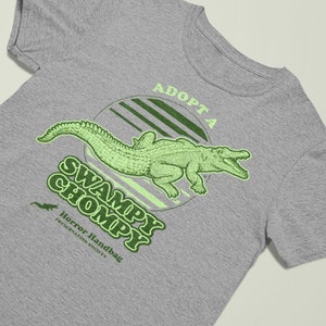 May include: A grey t-shirt with a green alligator graphic and the text "Adopt a Swampy Chompy" and "Horror Handbag Preservation Society".
