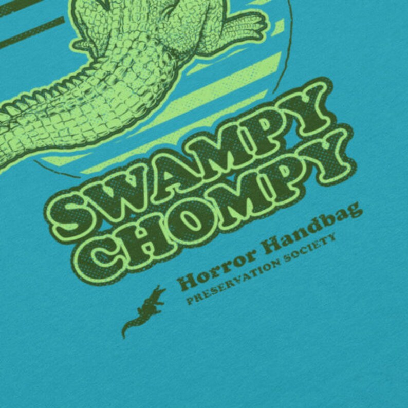 May include: A teal blue t-shirt featuring a green alligator graphic and the text "Swampy Chompy Horror Handbag Preservation Society".