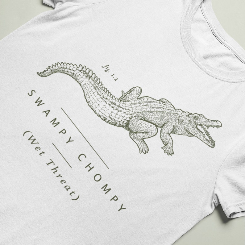 May include: White t-shirt featuring a detailed illustration of an alligator with its mouth open. The text "SWAMPY CHOMPY (Wet Threat)" and "fig. 1.2" are printed in a dark green color.