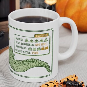 May include: White ceramic mug with a green alligator tail design. The mug has a black rim and handle. The mug has text that reads "DANGER: AAAA KIND TO OTHERS: NOT VERY HANDBAGS: CREDIT SCORE: POOR".