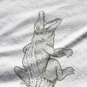 May include: A detailed illustration of an alligator in a sketch style. The alligator is depicted with its mouth open, revealing teeth, and its body is textured with scales. The image is in shades of gray and white.