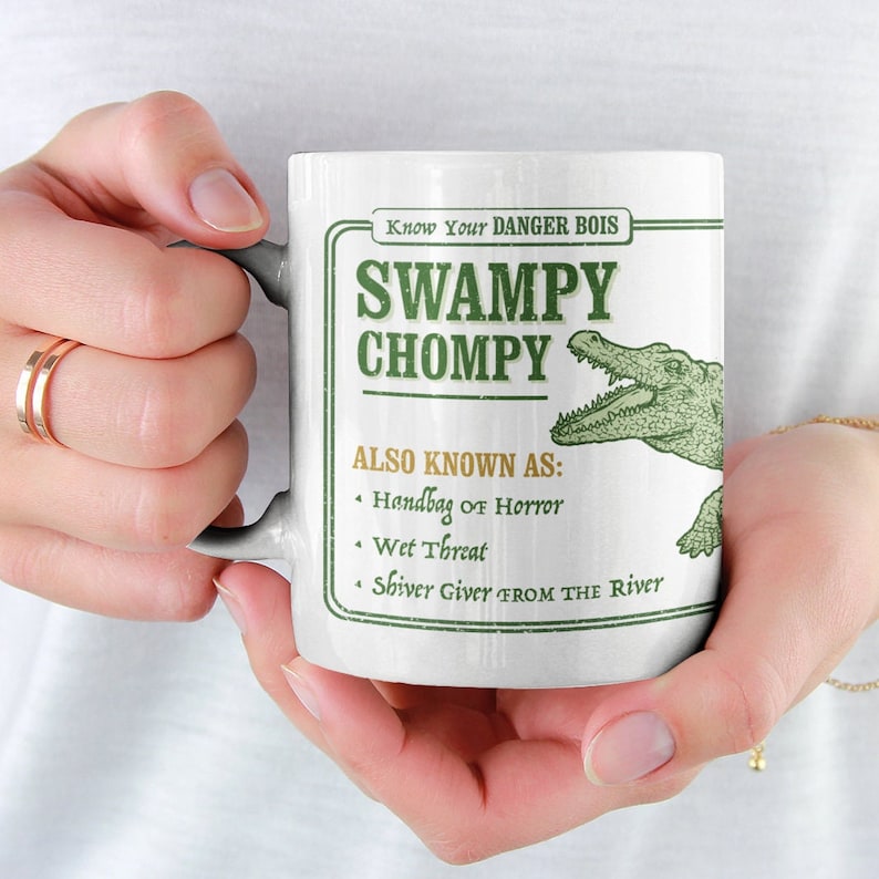 May include: White ceramic mug with a green alligator graphic and the text "Know Your DANGER BOIS SWAMPY CHOMPY ALSO KNOWN AS: Handbag of Horror Wet Threat Shiver Giver FROM THE River"