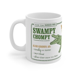 May include: White ceramic mug with a green graphic of an alligator with the text "Know Your DANGER BOIS SWAMPY CHOMPY ALSO KNOWN AS: Handbag of Horror Wet Threat Shiver Giver FROM THE River"