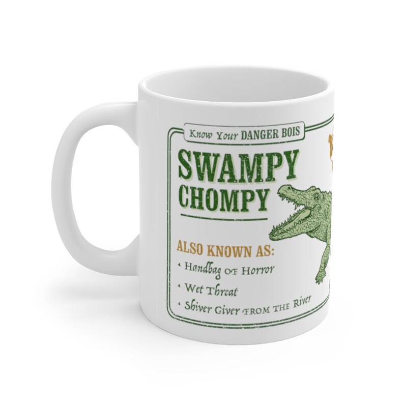 May include: White ceramic mug with a green graphic of an alligator with the text "Know Your DANGER BOIS SWAMPY CHOMPY ALSO KNOWN AS: Handbag of Horror Wet Threat Shiver Giver FROM THE River"