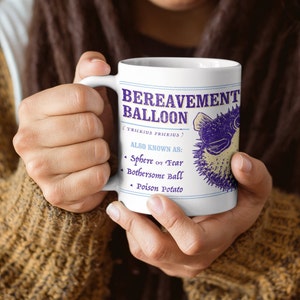 May include: White ceramic mug with a purple and white illustration of a pufferfish. The text on the mug reads "BEREAVEMENT BALLOON (TRICKIUS PRICKIUS) ALSO KNOWN AS: Sphere of Fear Bothersome Ball Poison Potato"