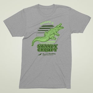 May include: A grey t-shirt featuring a green alligator graphic and the text "Adopt a Swampy Chompy" in a retro style. The text "Horror Handbag Preservation Society" is also included in smaller text below the graphic.