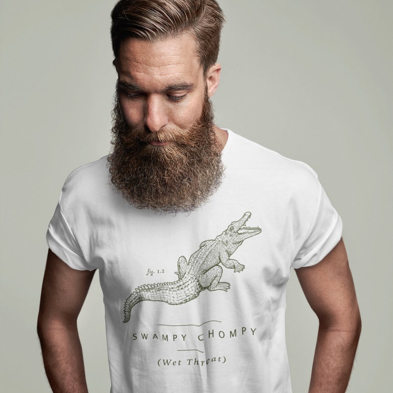 May include: White t-shirt featuring a detailed illustration of an alligator with the text "Swampy Chomp" and "(Wet Threat)" in a green font. The graphic is a vintage style design.