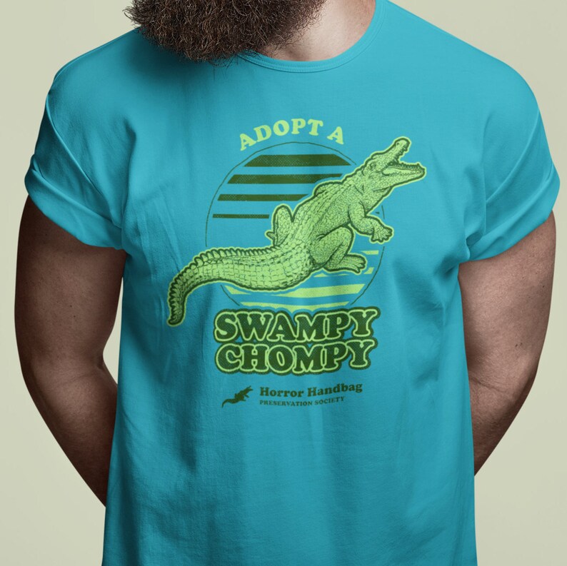 May include: A turquoise blue t-shirt featuring a green alligator graphic and the text "Adopt a Swampy Chompy" in a retro font. The text "Horror Handbag Preservation Society" is also included in smaller font.