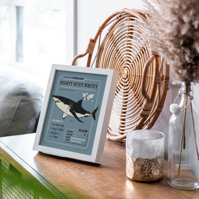 May include: A white framed print with a blue background featuring a black and white illustration of a great white shark. The print has the title "Mighty Bitey Whitey" and includes information about the shark, such as its scientific name and danger level.