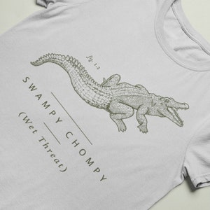 May include: A light gray t-shirt featuring a detailed illustration of an alligator in a vintage style. The text "SWAMPY CHOMPY (Wet Threat)" is printed below the alligator. The shirt has a classic crew neck.