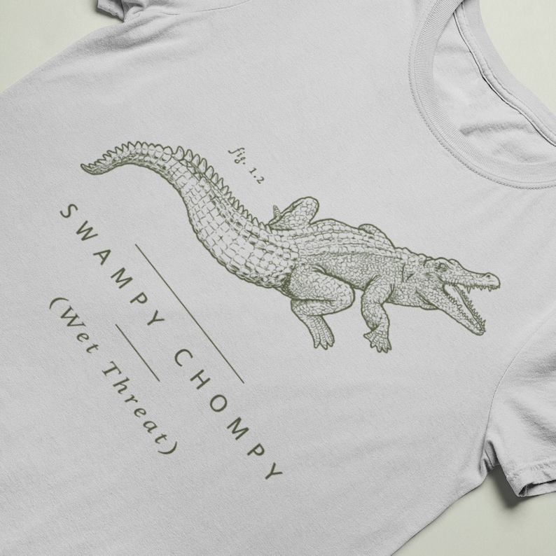 May include: A light gray t-shirt featuring a detailed illustration of an alligator in a vintage style. The text "SWAMPY CHOMPY (Wet Threat)" is printed below the alligator. The shirt has a classic crew neck.
