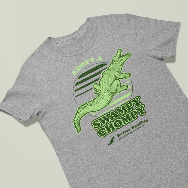 May include: A grey t-shirt featuring a green alligator graphic and the text "Adopt a Swampy Chompy Horror Handbag Preservation Society".